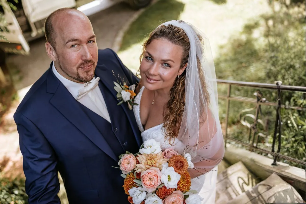 The bride and groom look at the camera together, holding a bouquet and wearing elegant wedding attire in Schloss Marbach.